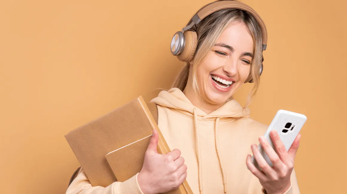 Młoda kobieta w beżowej bluzie i słuchawkach na uszach trzyma książki oraz smartfon, uśmiechając się do ekranu na tle w ciepłym odcieniu beżu.